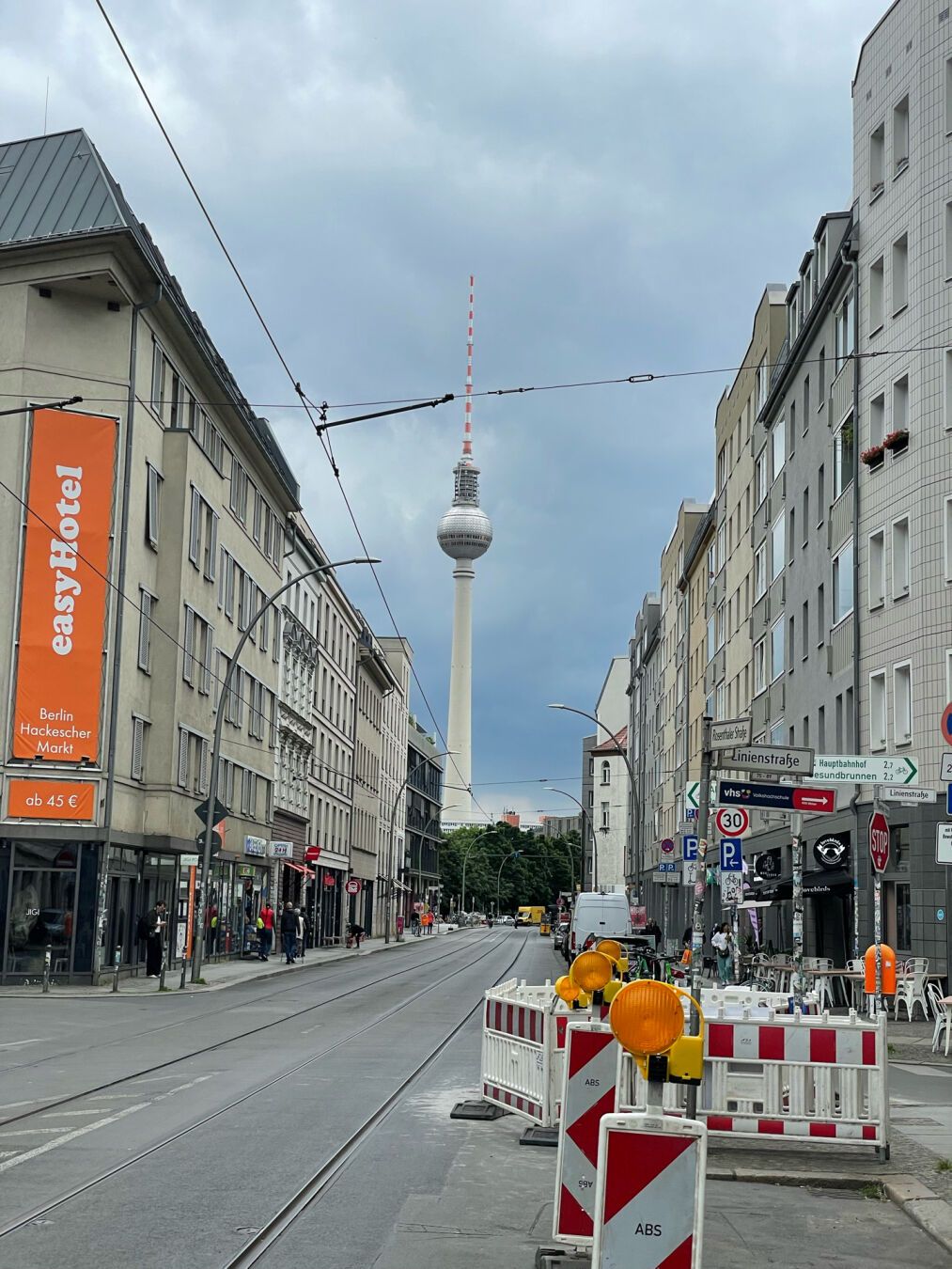 In the photo you can see a street, and at the end of it, you can see the tower in Alexander Platz (Berlin). The sky is cloudy.