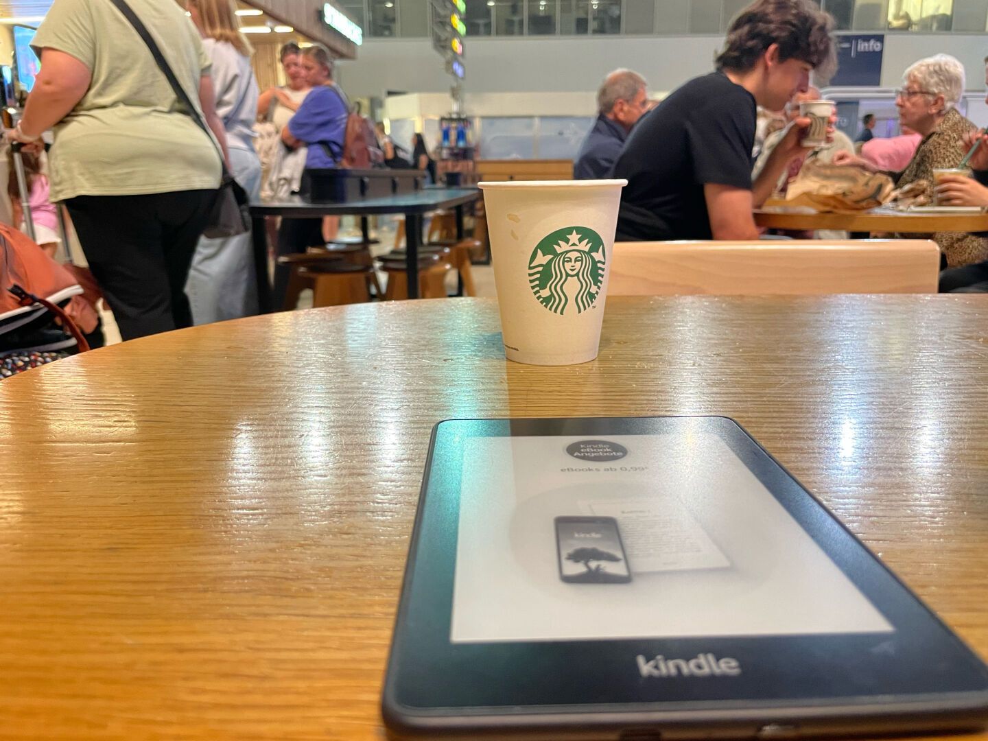 In the photo you can see a Starbucks coffee and a Kindle on a table, and some people in the background lining up to get their coffee. We are at the Alicante airport waiting to board our flight.